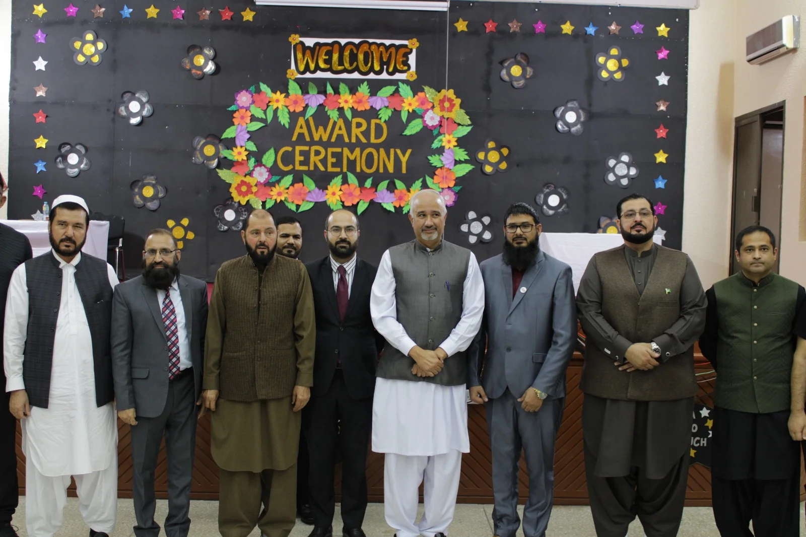 A group of men standing together in front of a decorated Award Ceremony backdrop, with fellow members and representatives of the Pakistani Islamic Private School Al Ain community.